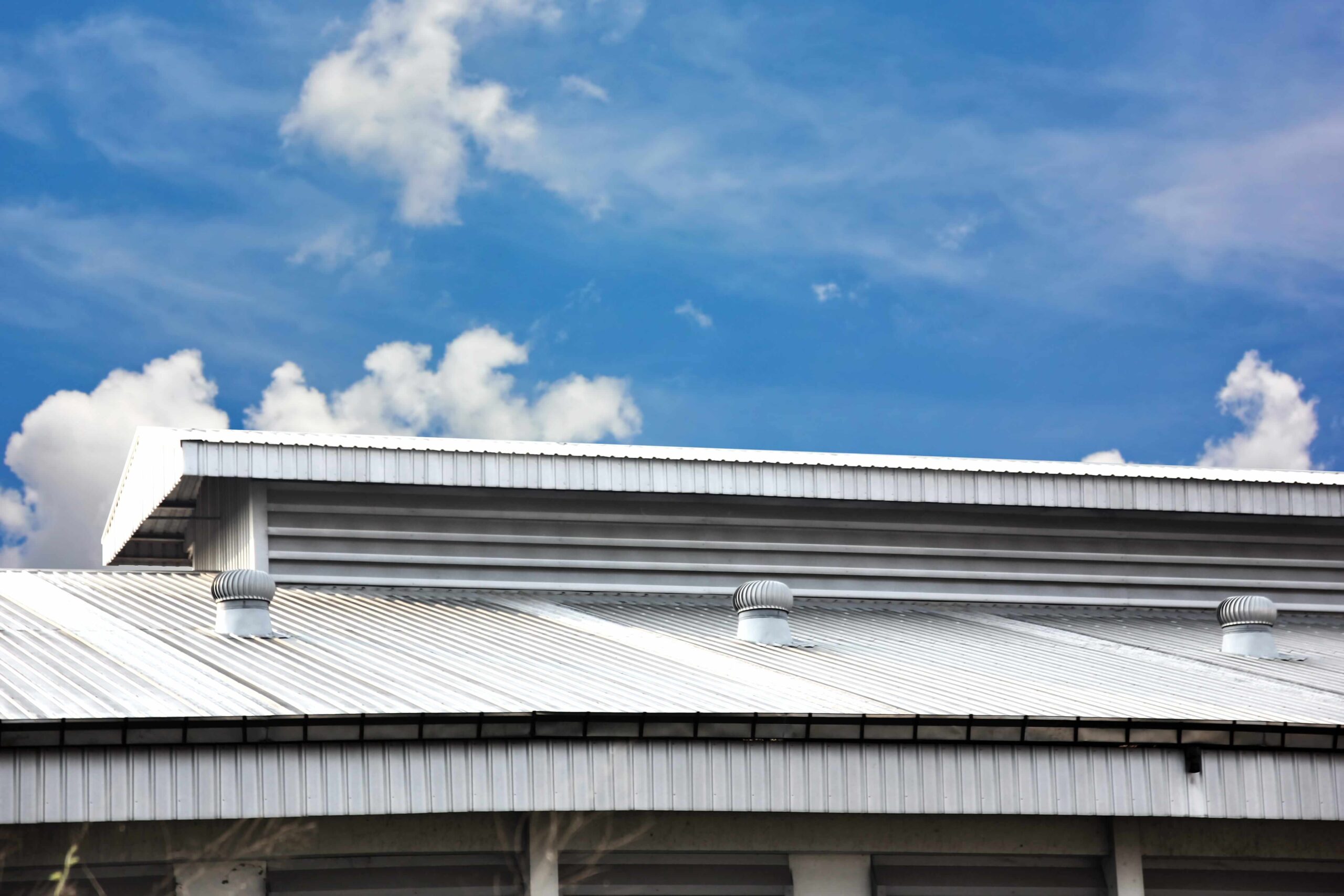 Metal roofing on a commercial warehouse with ventilation turbines under a blue sky with clouds.